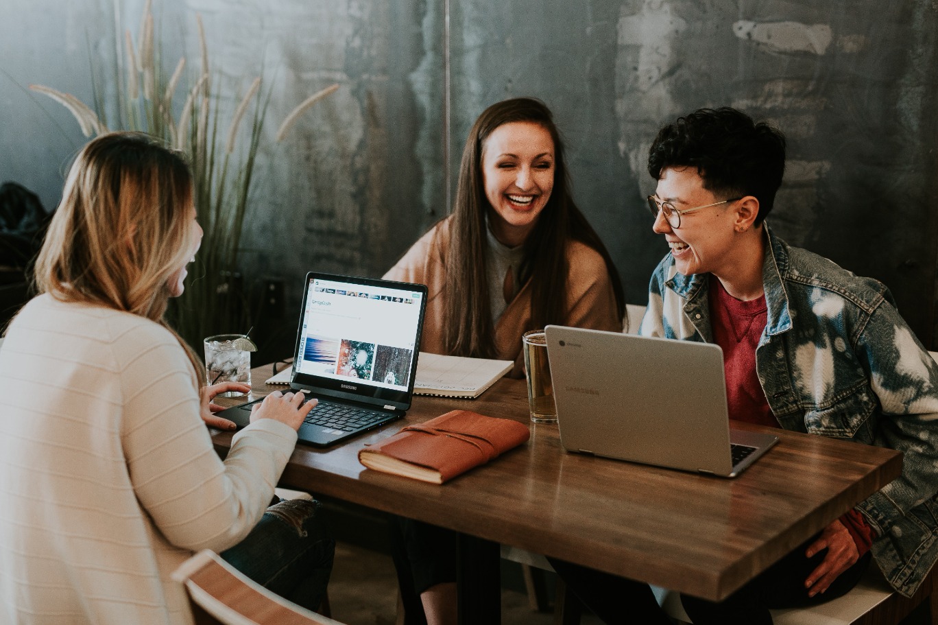 Trois personnes sont assises à une table en bois, chacune avec un ordinateur portable. Elles sourient et semblent avoir une conversation agréable. Le cadre est décontracté et détendu, avec une plante visible en arrière-plan.