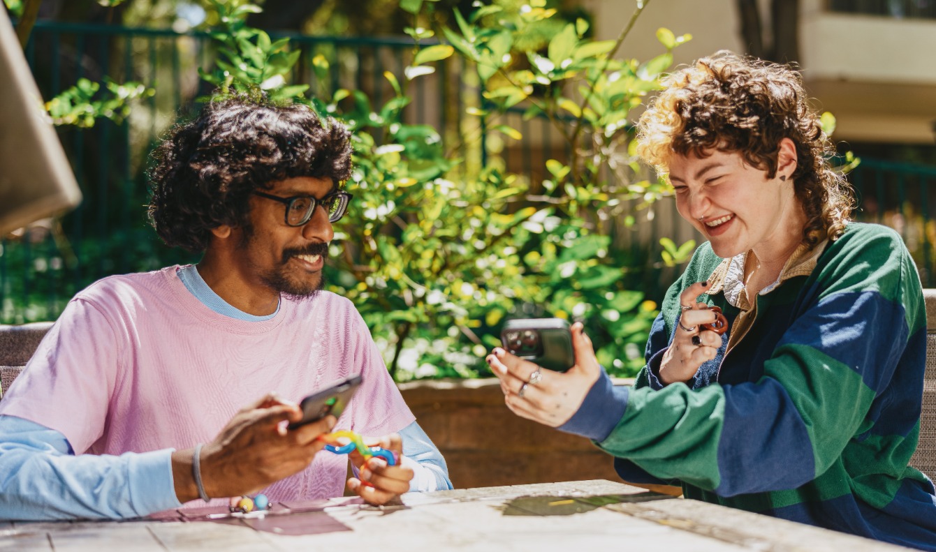 Deux personnes souriantes regardent leur smartphone assises à une table en plein air. La personne de gauche a les cheveux bouclés et des lunettes, porte une chemise rose. La personne de droite a les cheveux courts et bouclés et une veste multicolore. Une végétation luxuriante en arrière-plan.