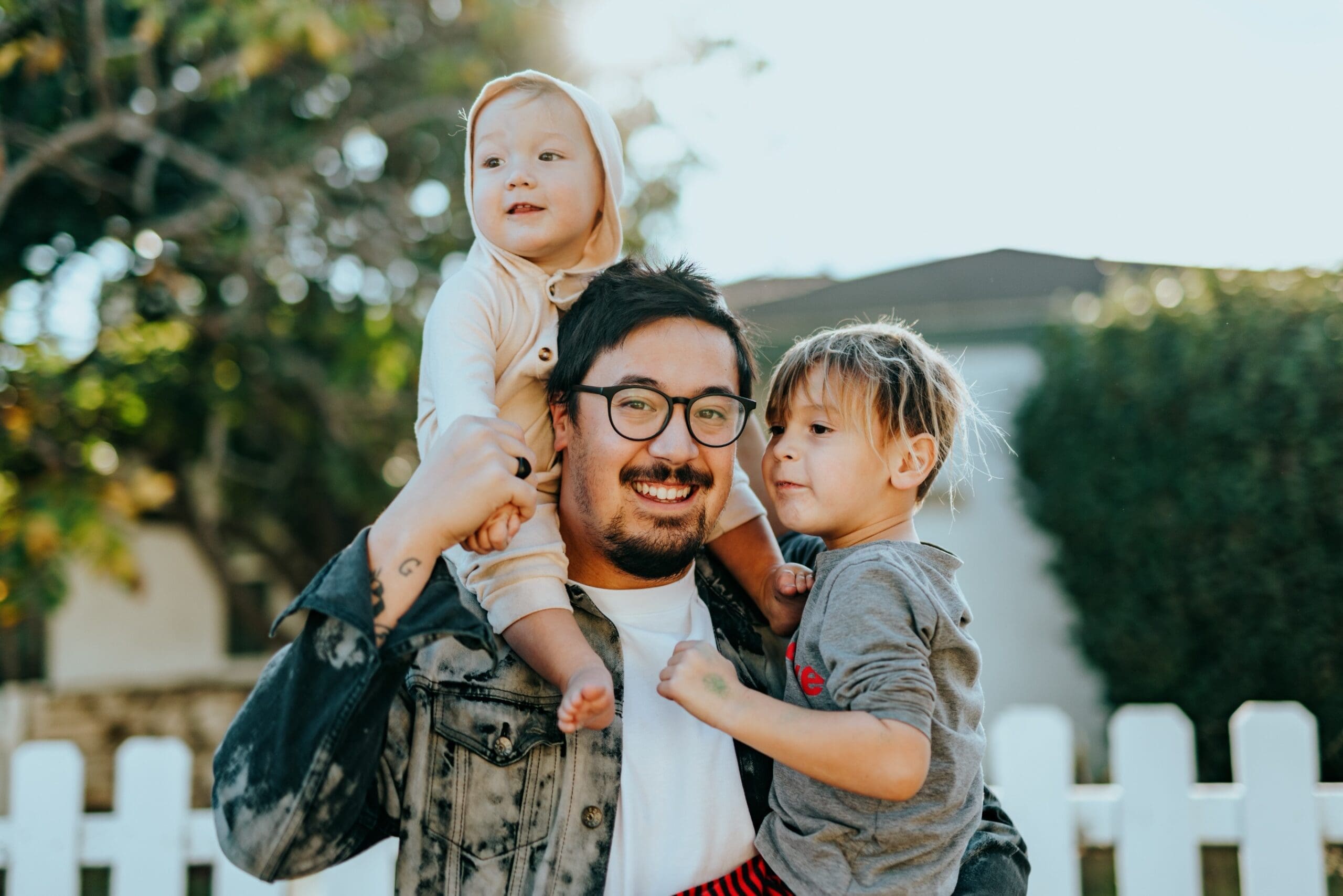 Un homme souriant portant des lunettes tient deux jeunes enfants à l'extérieur, près d'une clôture blanche et d'un espace vert. L'un des enfants est assis sur ses épaules, vêtu d'une tenue de couleur claire, tandis que l'autre se tient à côté de lui, vêtu d'un haut gris.