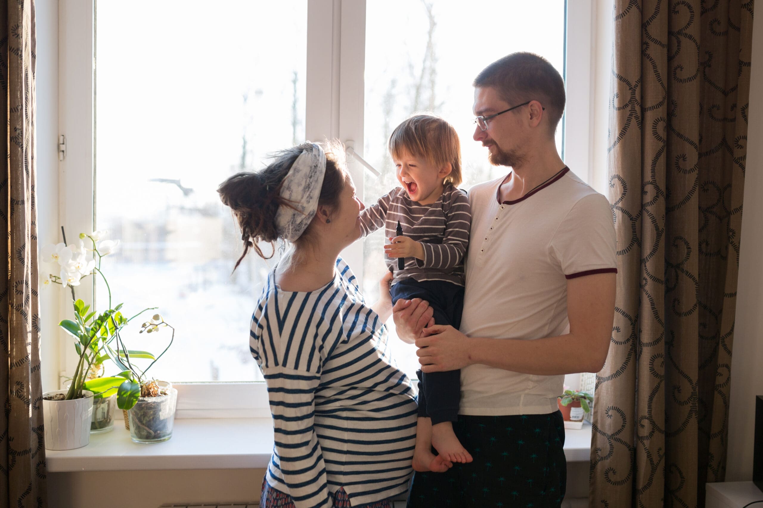 Une scène familiale joyeuse devant une fenêtre. Une femme enceinte en chemise rayée sourit à un homme portant des lunettes tenant un bambin qui rit. La lumière du soleil filtre à travers la fenêtre, illuminant leurs visages et trois plantes en pot sur le rebord de la fenêtre.
