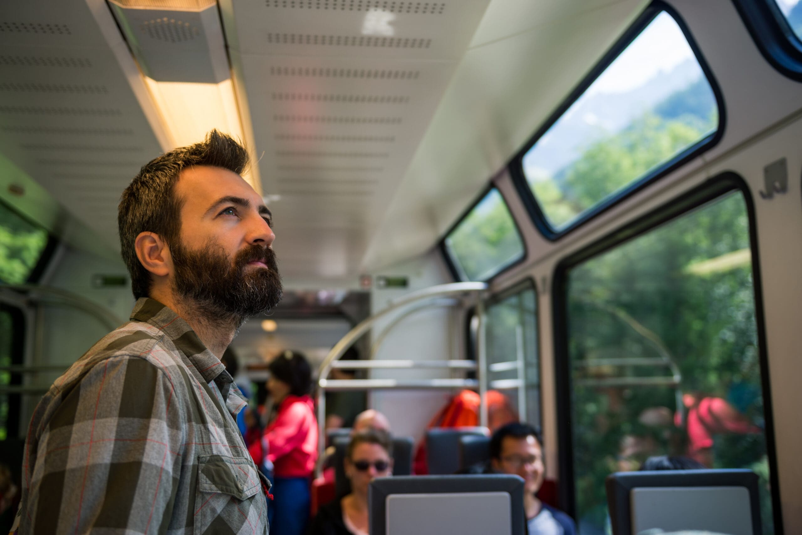 Un homme barbu portant une chemise à carreaux se tient debout dans un wagon et regarde par les grandes fenêtres. D'autres passagers sont assis à proximité. La vue extérieure montre de la verdure et un paysage montagneux sous un ciel clair.