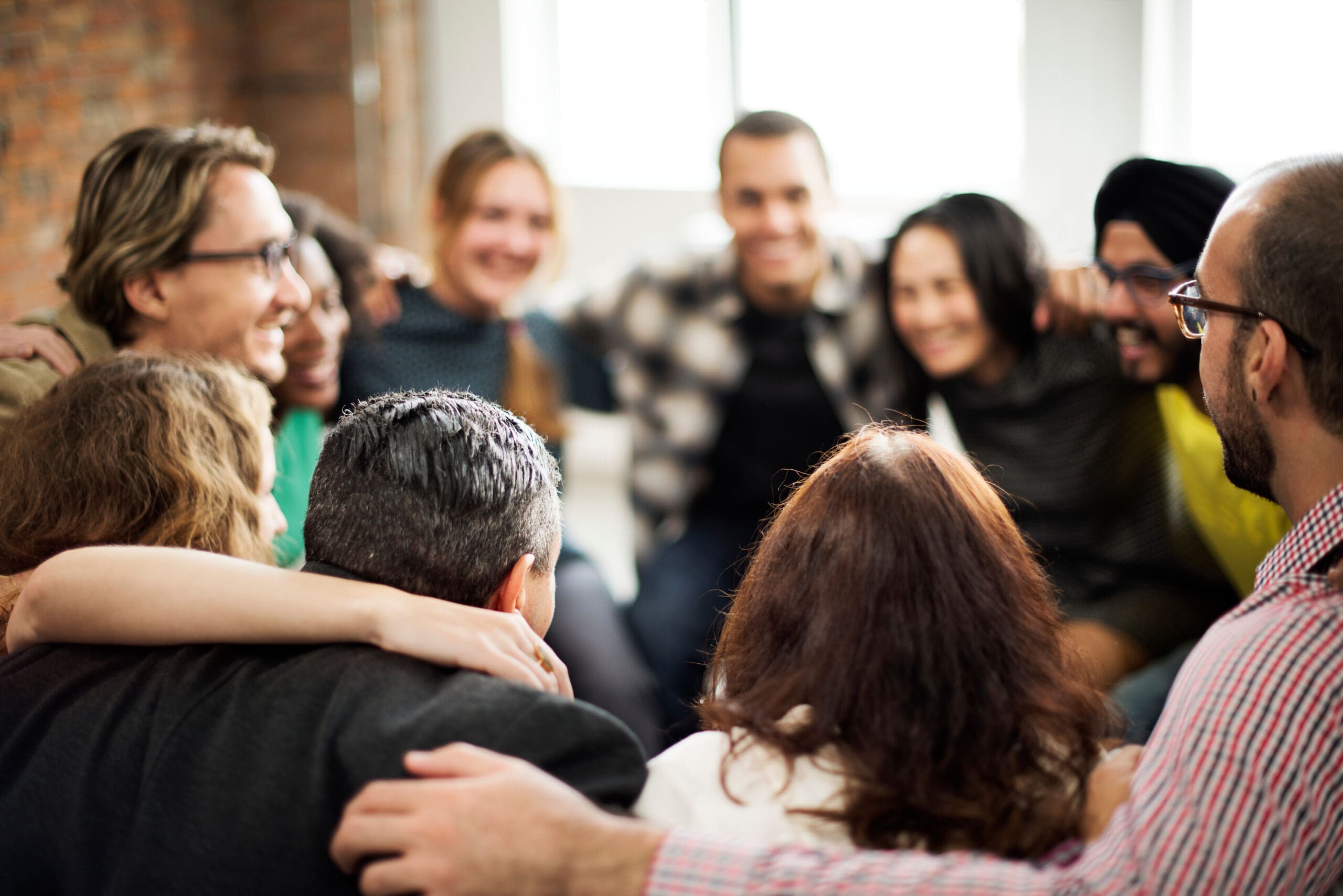 Un groupe diversifié de personnes assises en cercle, souriantes et s'enlaçant les unes les autres, transmettant un sentiment d'unité et de travail d'équipe. L'arrière-plan est légèrement flou, soulignant leur lien.