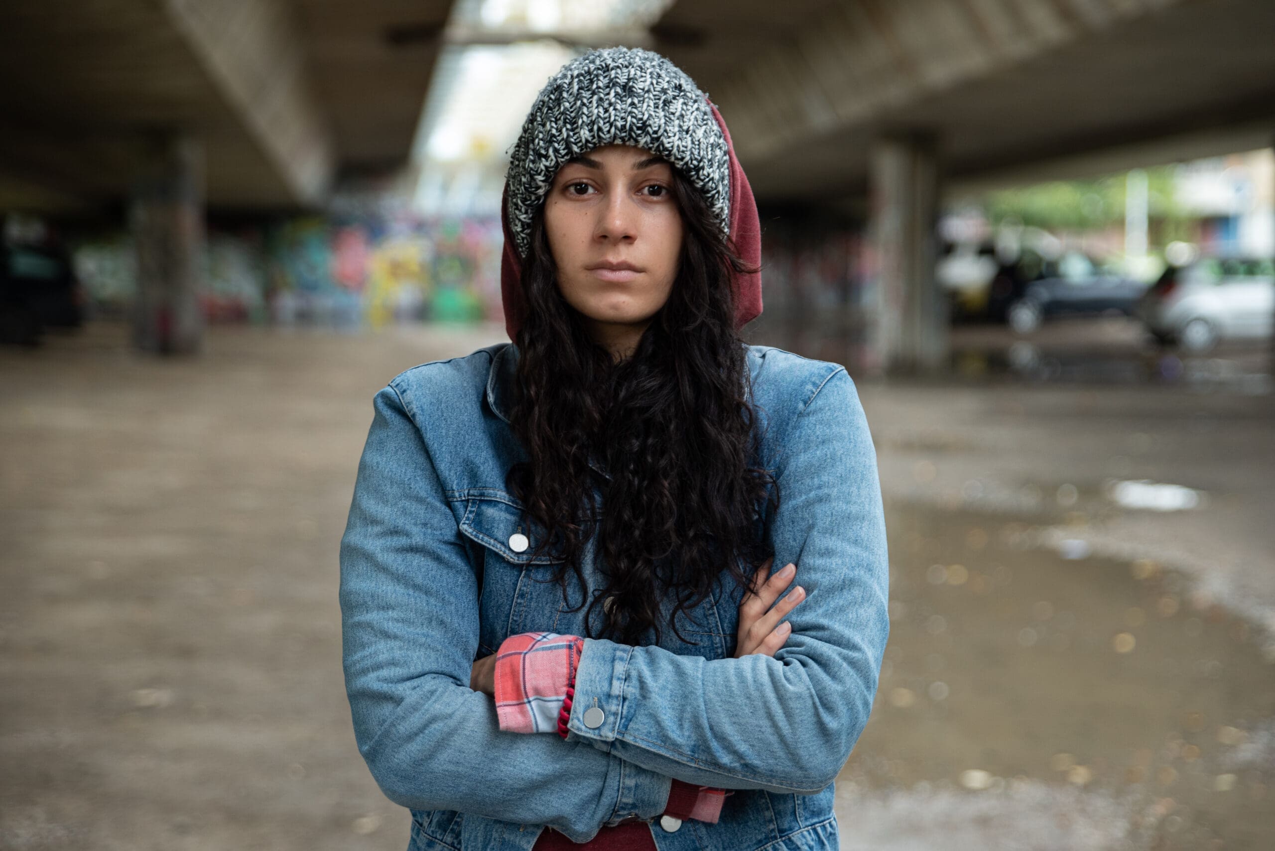 Une personne aux cheveux longs et noirs se tient debout sous un pont, vêtue d'une veste en jean, d'une chemise à carreaux et d'un bonnet en tricot. Son expression est sérieuse et ses bras sont croisés. L'arrière-plan est flou, avec des murs couverts de graffitis et un sol humide.