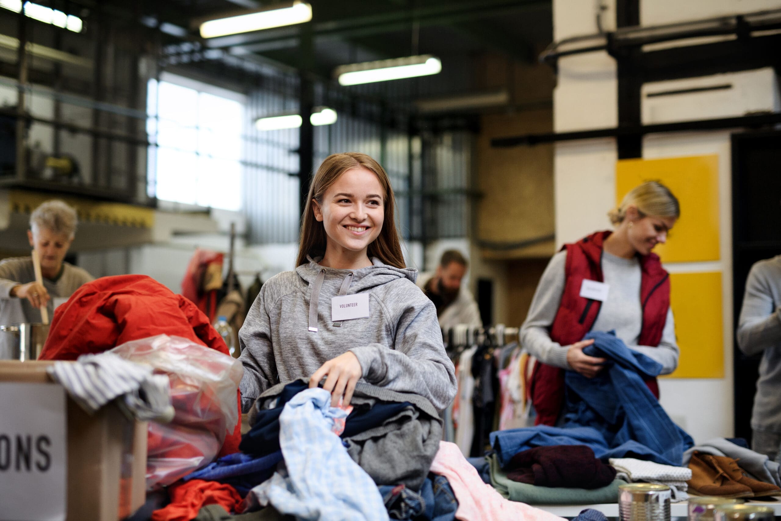 Une jeune femme sourit en triant des vêtements dans un centre de dons. Elle est entourée de sacs de vêtements sur une table. D'autres bénévoles à l'arrière-plan sont occupés à organiser et à plier des vêtements.