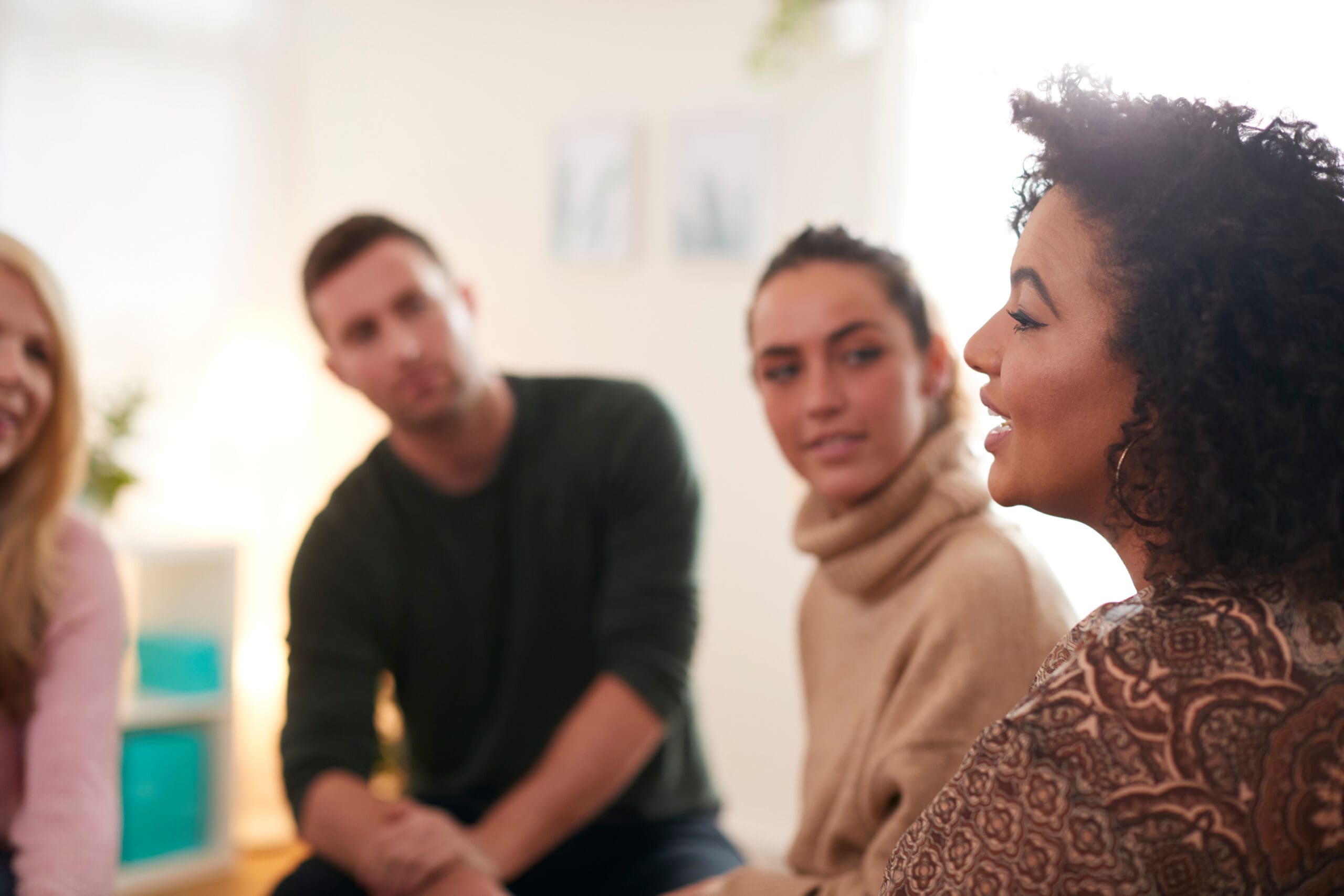 Un groupe de quatre personnes assises en cercle, en pleine conversation. L'accent est mis sur une femme aux cheveux bouclés qui parle, tandis que les autres écoutent attentivement. Le décor semble être une pièce décontractée et bien éclairée.