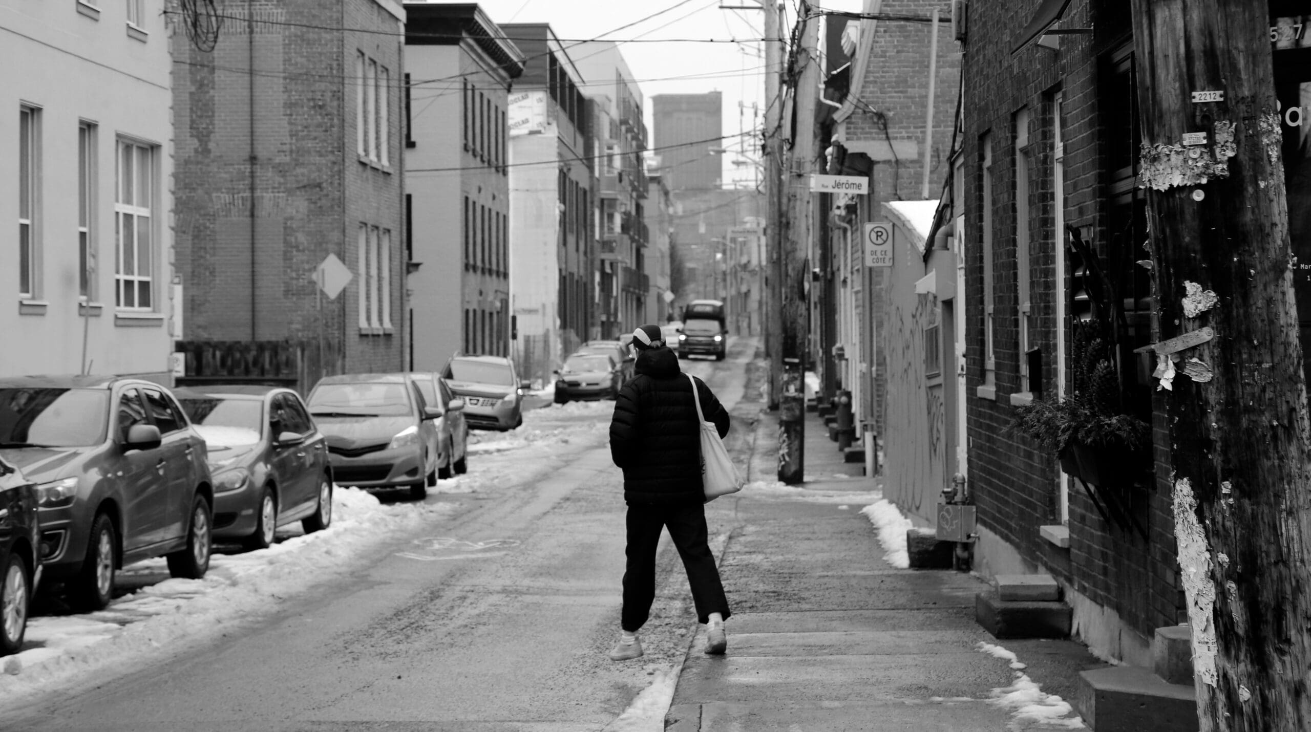 Une personne portant une veste à capuche marche dans une rue étroite et enneigée, bordée de voitures garées et de bâtiments en briques. Des câbles électriques traversent le ciel nuageux et des plaques de neige recouvrent le trottoir et la route.