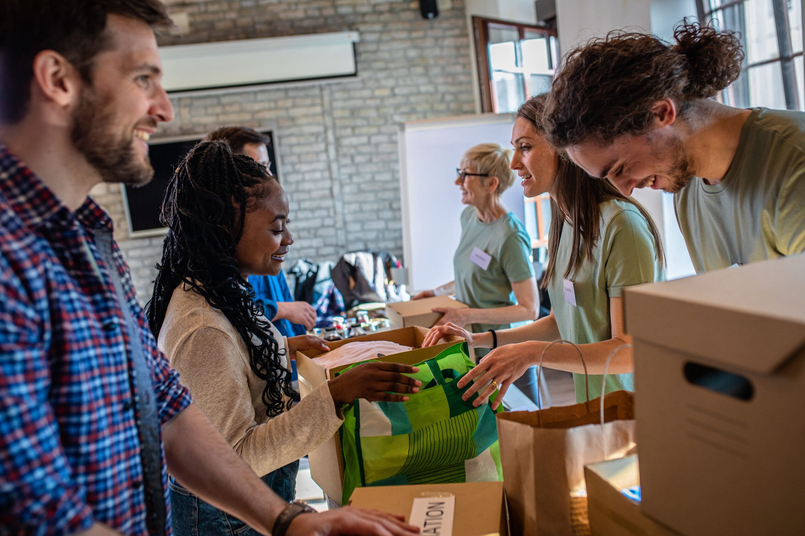 Un groupe de personnes se tient autour d'une table à l'intérieur, triant et emballant des objets dans des sacs et des cartons. Ils semblent engagés et joyeux, participant à ce qui semble être un service communautaire ou une activité bénévole.