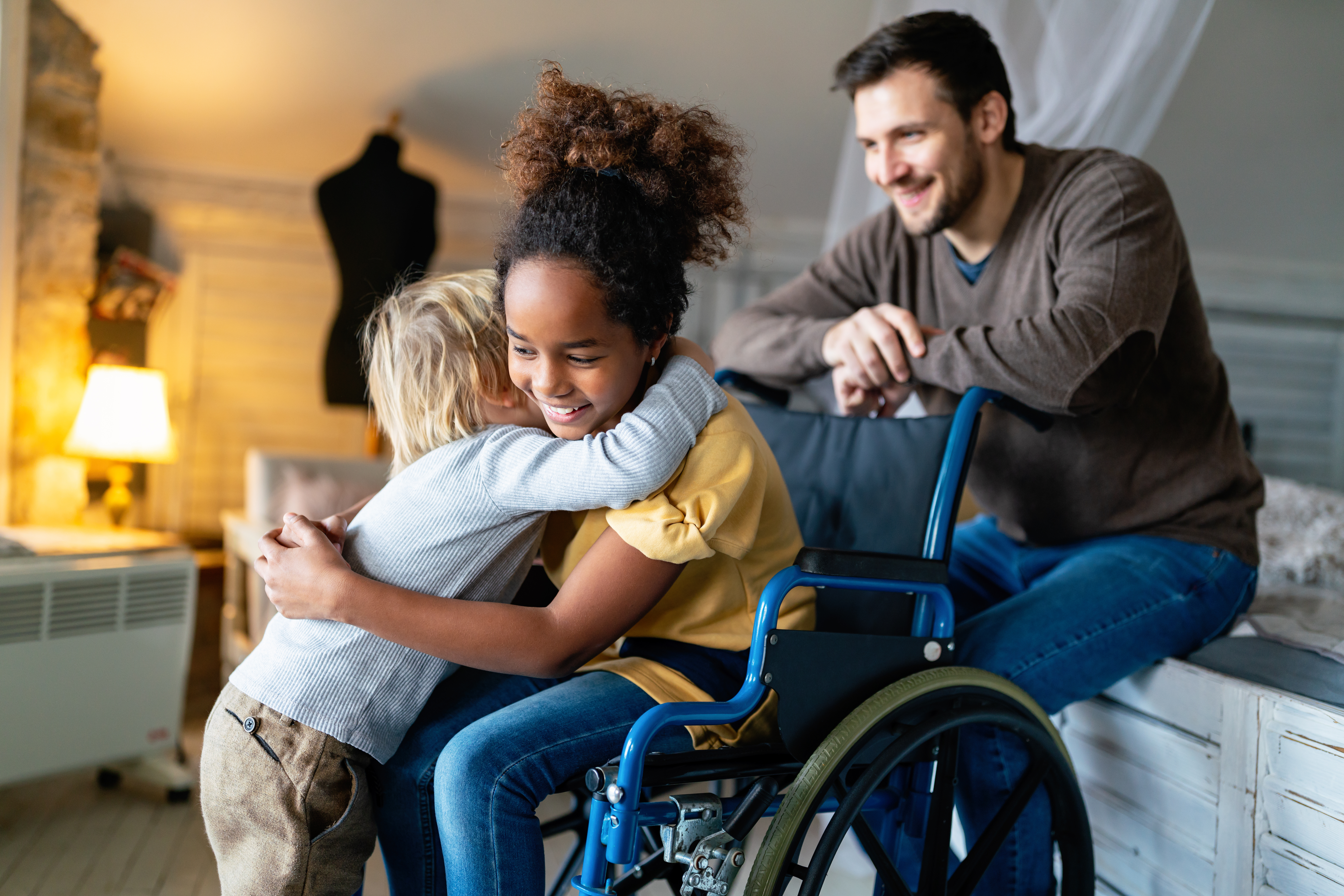 Un enfant en chemise jaune est assis dans un fauteuil roulant, souriant tout en serrant dans ses bras un autre enfant aux cheveux blonds. Un homme en pull marron sourit et les regarde de près. Le décor semble être une pièce confortable avec un éclairage chaleureux.