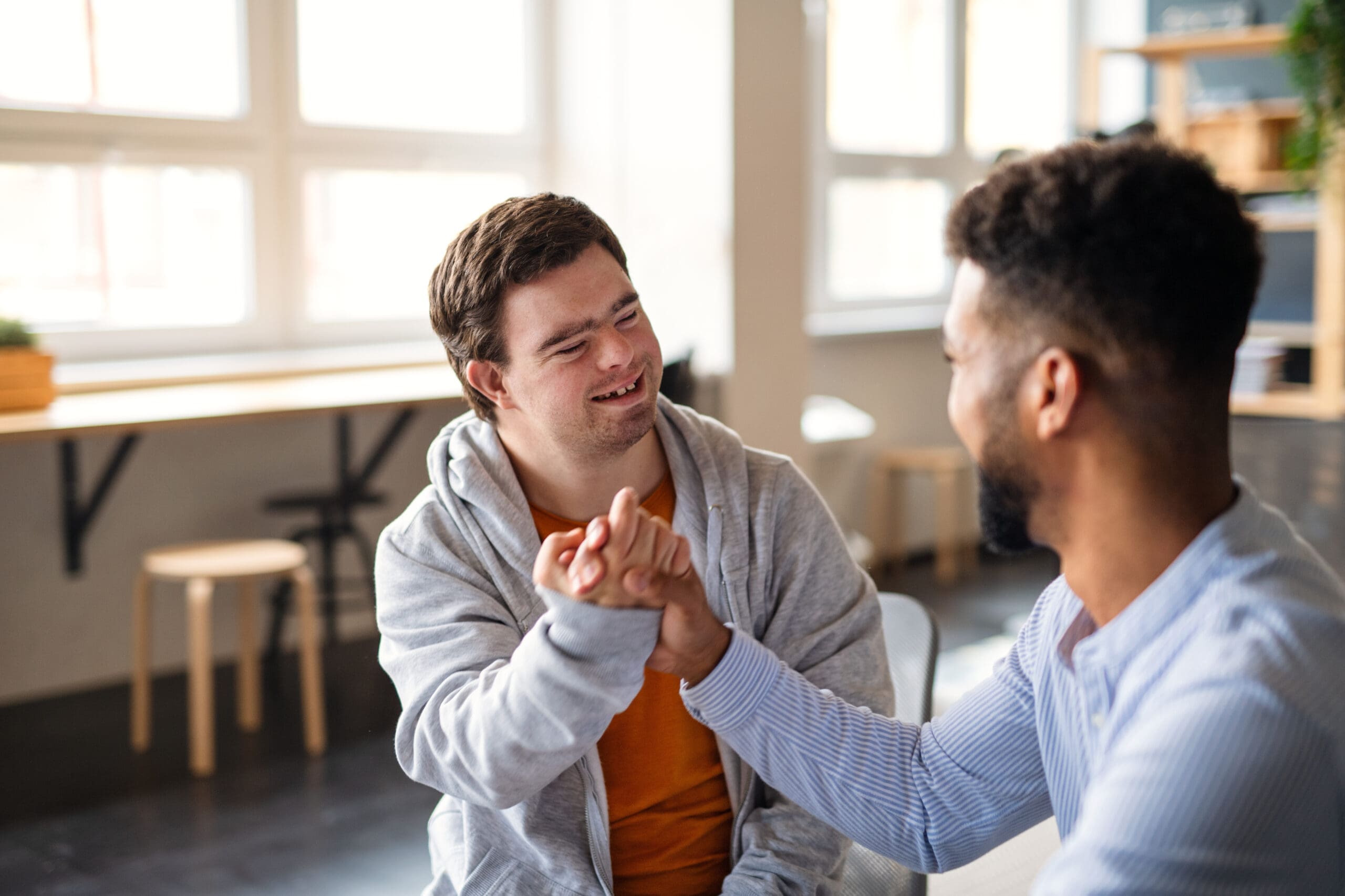 Deux hommes souriants et assis à l'intérieur, se donnant un coup de poing. L'un porte un sweat à capuche gris et l'autre, avec une barbe, porte une chemise rayée. L'arrière-plan présente de grandes fenêtres et des meubles en bois.