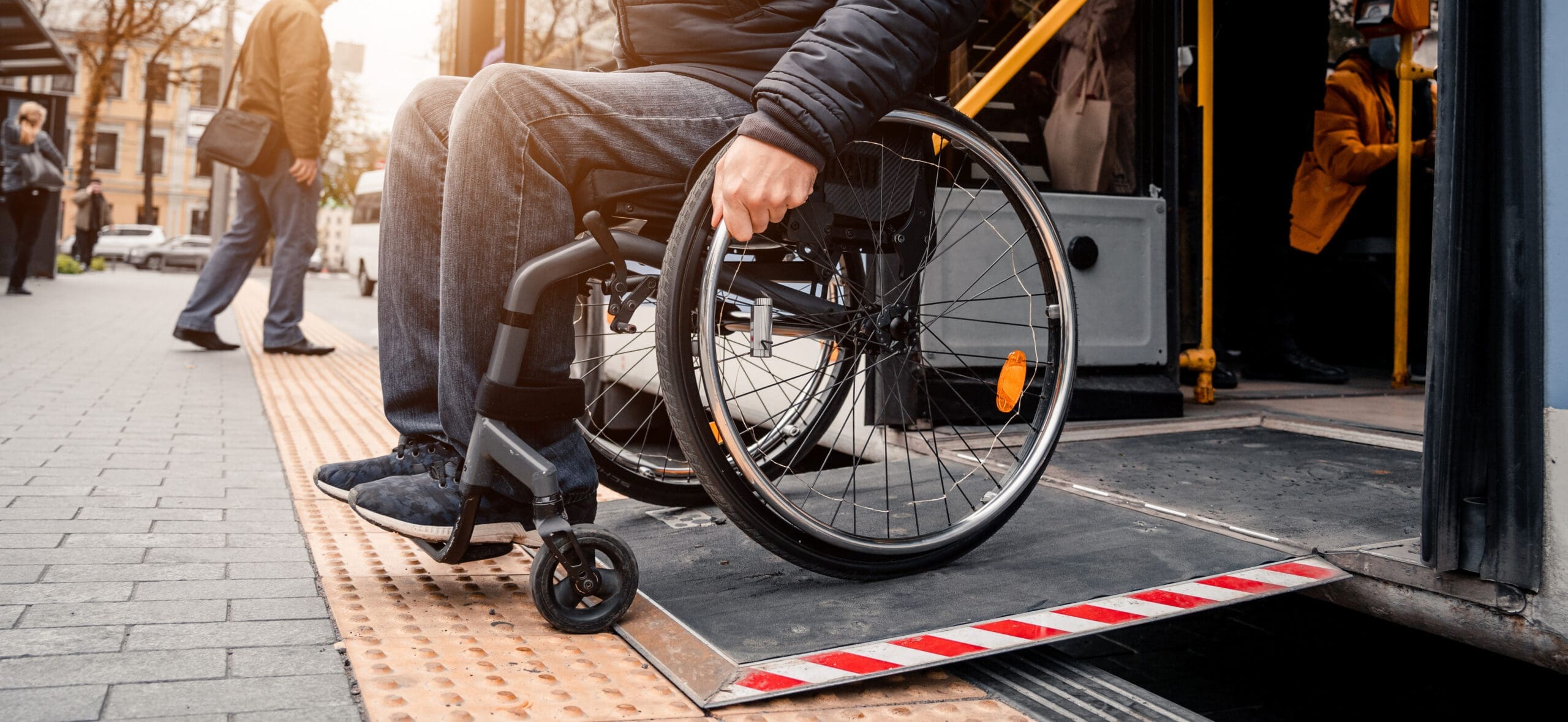 Une personne en fauteuil roulant utilise une rampe pour monter dans un bus public. La scène se déroule dans une rue de la ville avec des gens qui marchent en arrière-plan. Le soleil se couche, projetant une douce lueur.