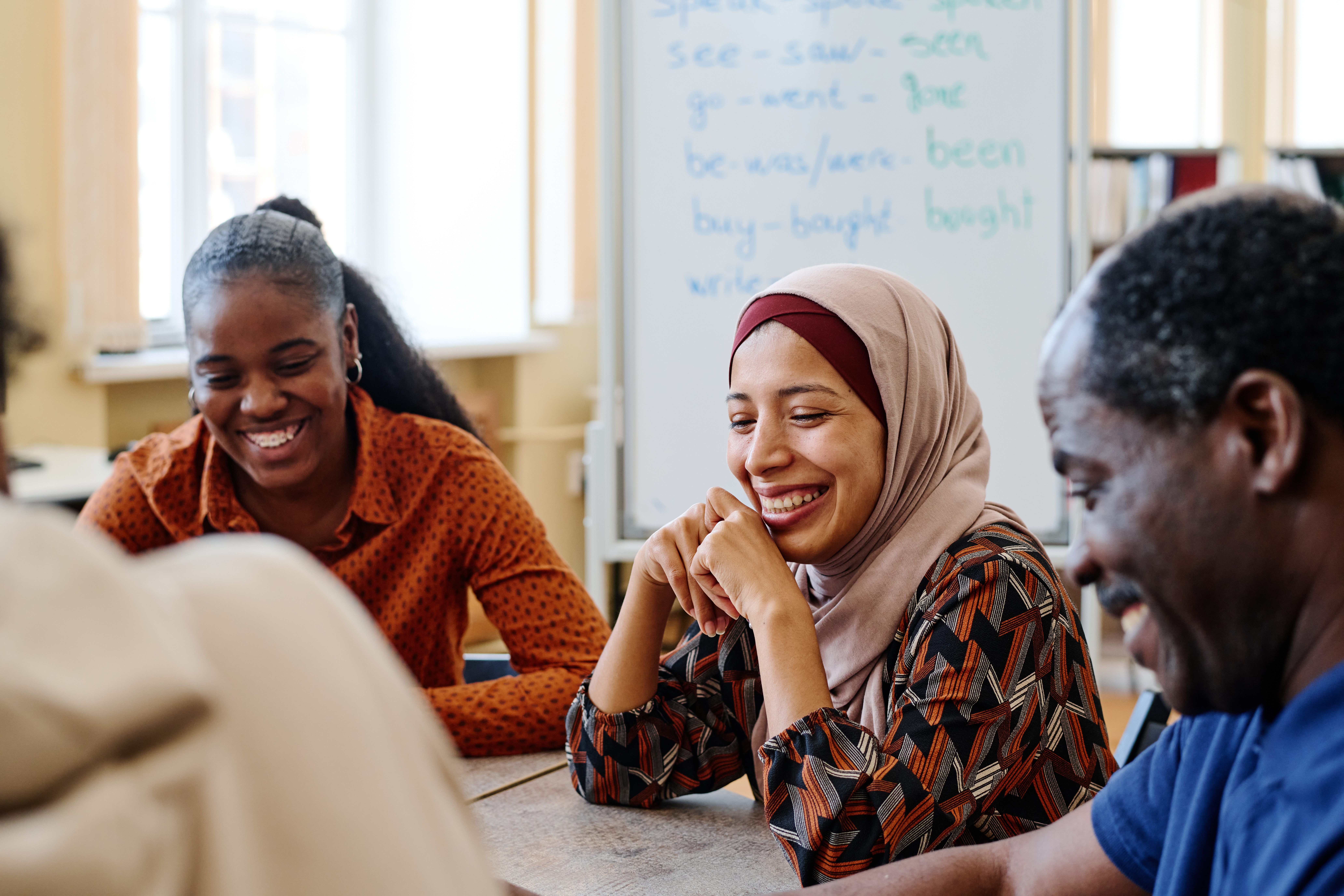 Un groupe diversifié de personnes est assis autour d'une table, souriant et engagé dans une conversation. Il y a un tableau blanc avec des verbes anglais écrits en arrière-plan. Le décor semble être une salle de classe ou de réunion.