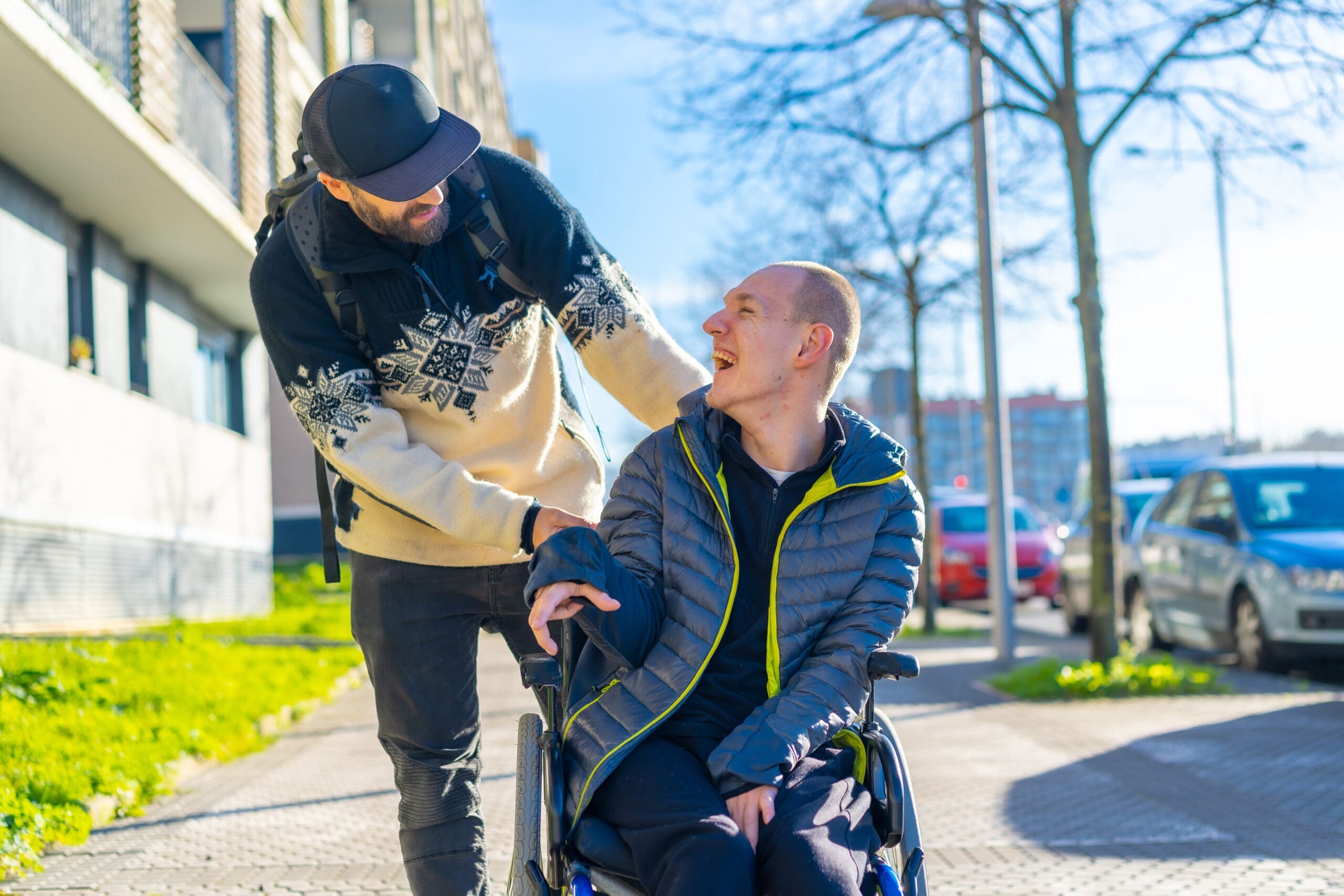 Un homme portant une casquette et un pull pousse un autre homme en fauteuil roulant sur un trottoir ensoleillé. Tous deux sourient et semblent profiter de leur temps libre. Des arbres et des voitures garées bordent la rue.