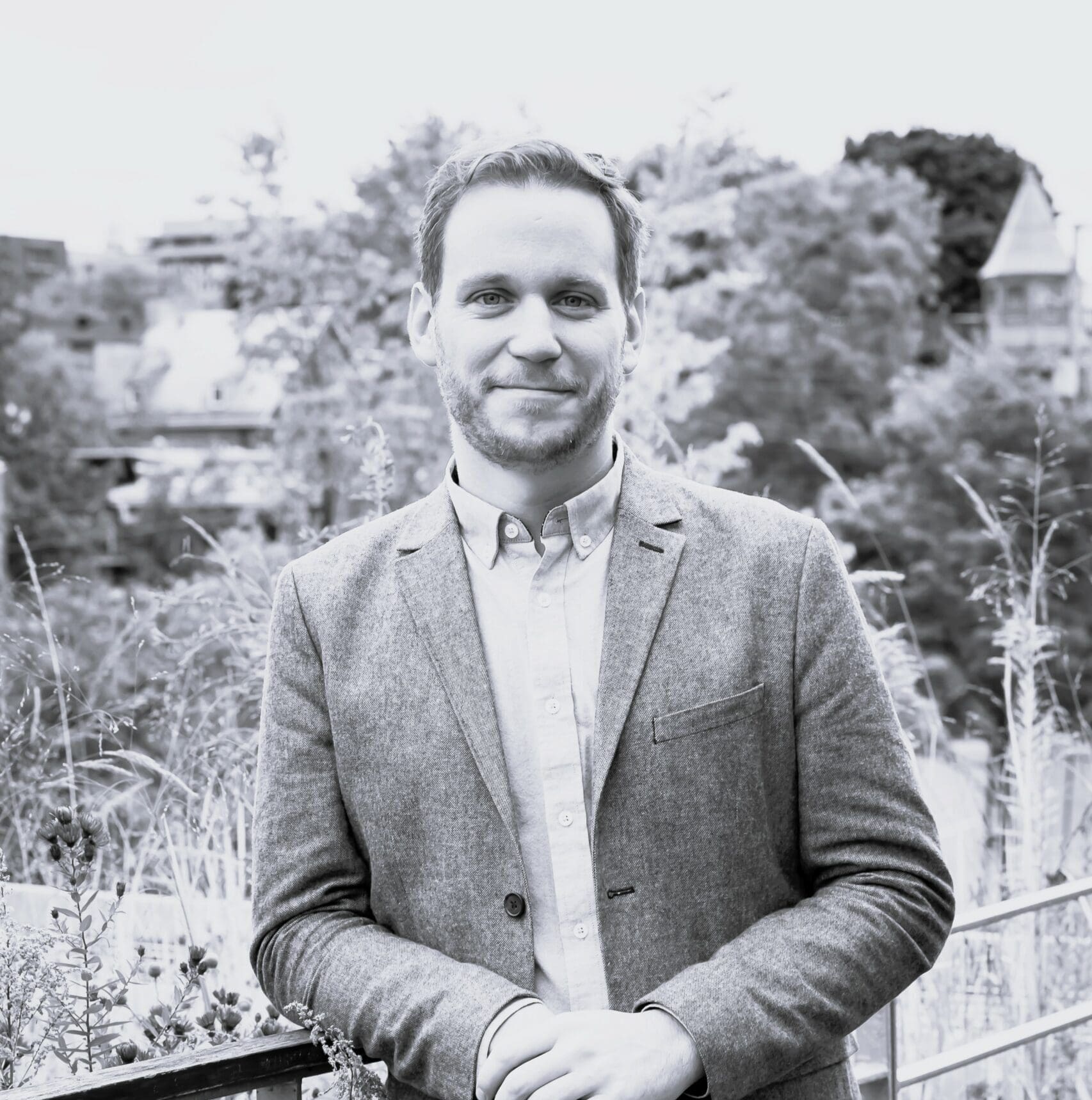 Un homme en blazer et chemise se tient dehors devant un paysage pittoresque composé d'arbres et de bâtiments. Il sourit, les mains posées sur une balustrade. L'image est en noir et blanc.