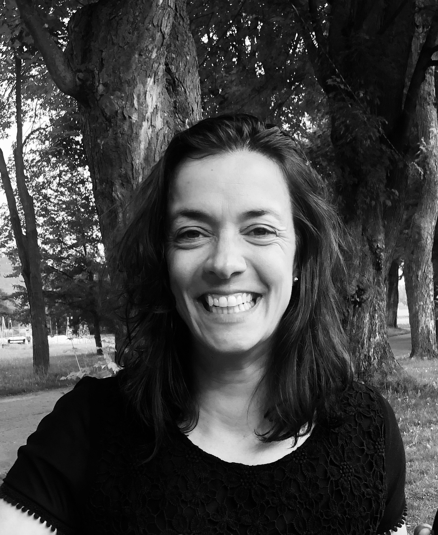 Une femme, souriant largement, se tient dehors devant un grand arbre. Elle porte un haut noir et a les cheveux noirs jusqu'aux épaules. L'arrière-plan est un décor de parc avec des arbres et un sentier. L'image est en noir et blanc.