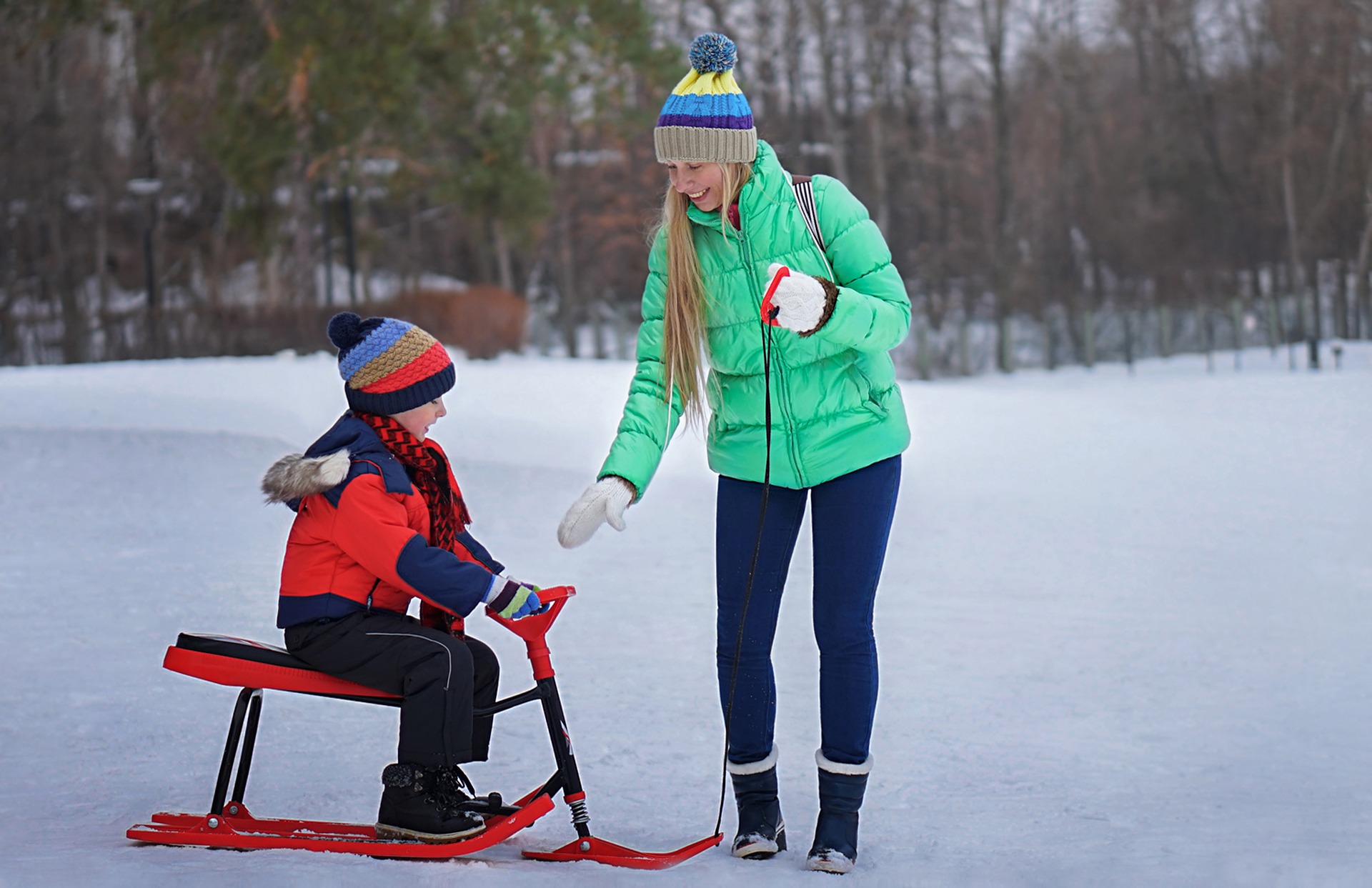 Une femme portant une veste verte et un chapeau bleu aide un enfant sur une luge rouge dans un parc enneigé. L'enfant, vêtu d'une tenue et d'un chapeau rouges et bleus, se prépare à faire de la luge sur la neige. Des arbres sont visibles en arrière-plan.