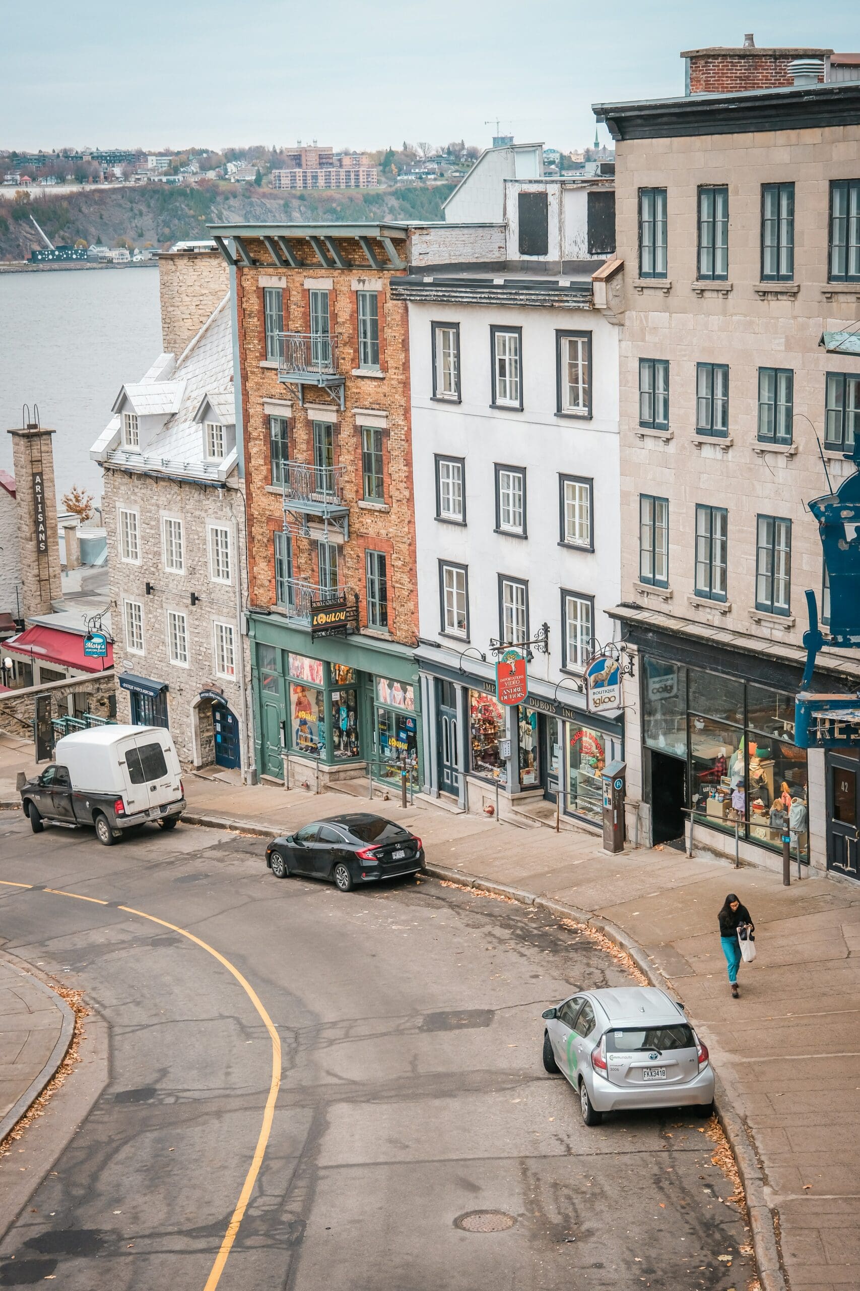 Rue bordée de bâtiments historiques, avec boutiques et cafés. Des voitures sont garées sur le côté et quelques piétons marchent. Surplombant une rivière et un paysage lointain sous un ciel couvert.