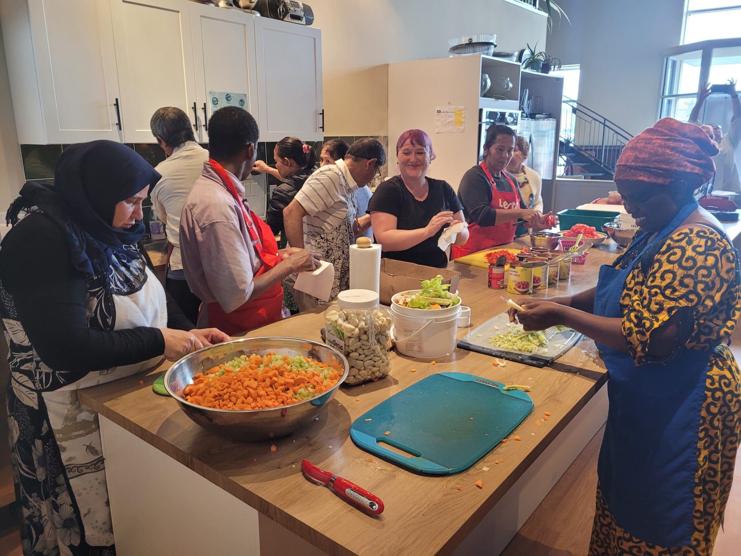 Un groupe diversifié de personnes se rassemble dans une cuisine, préparant ensemble de la nourriture. Ils coupent des légumes et portent des tabliers. La cuisine est spacieuse et bien éclairée, avec des ustensiles de cuisine et des ingrédients étalés sur le comptoir.
