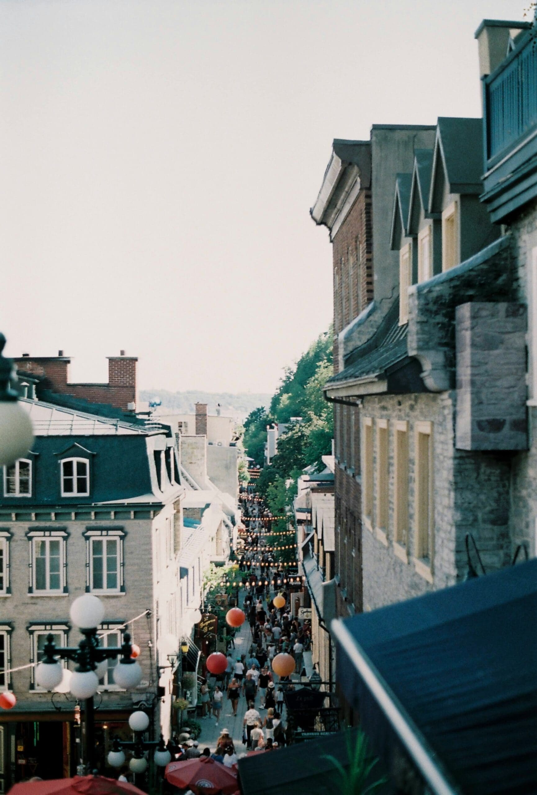 Une rue étroite dans une ville pittoresque bordée de bâtiments historiques. La rue est animée et décorée de lanternes en papier colorées. Des arbres et d'autres bâtiments sont visibles en arrière-plan sous un ciel clair.