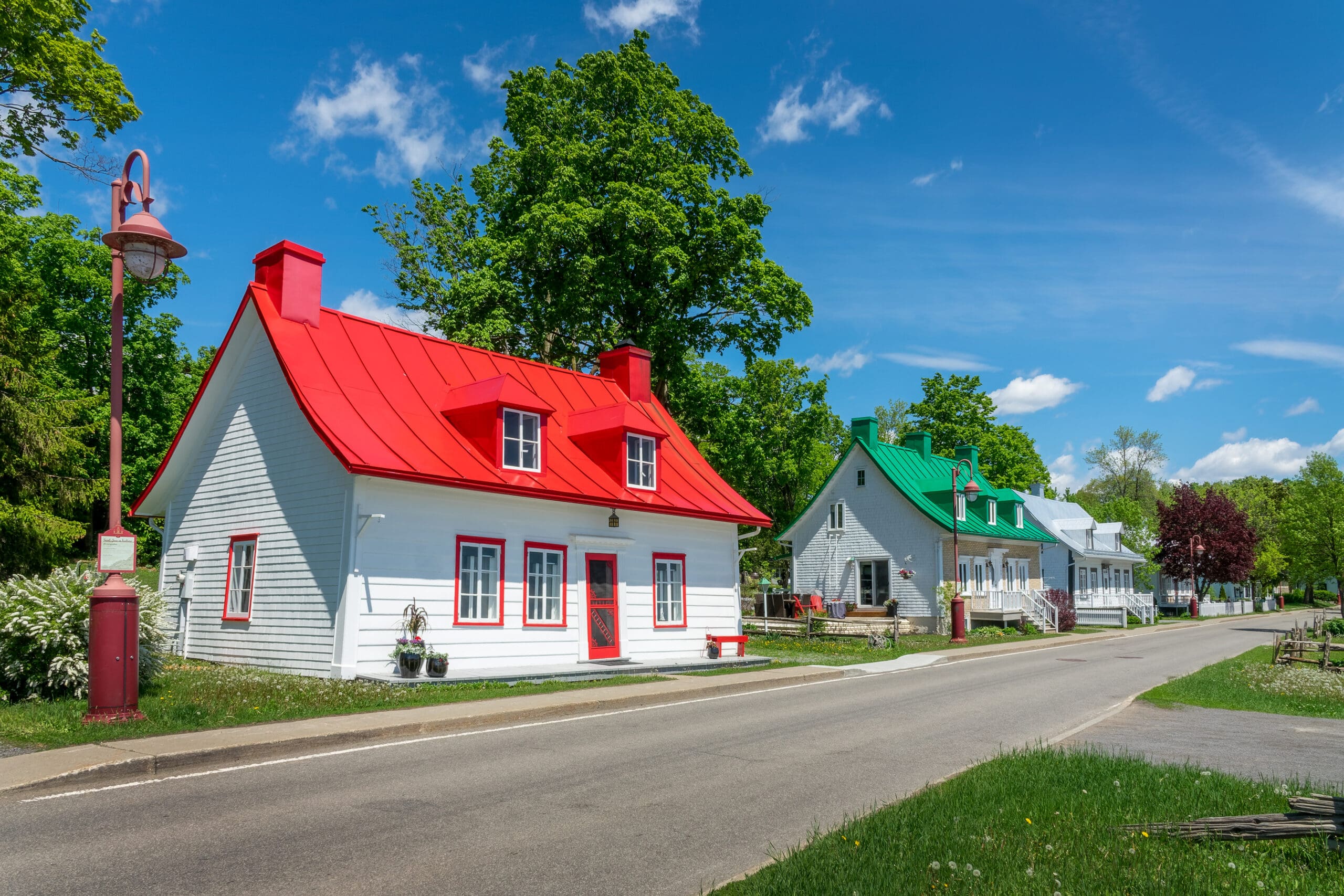 Une scène de rue pittoresque présente de charmantes maisons aux toits rouges et verts éclatants, sur fond d'arbres verts luxuriants et d'un ciel bleu vif. Un lampadaire rouge se dresse sur le trottoir, ajoutant au cadre pittoresque.