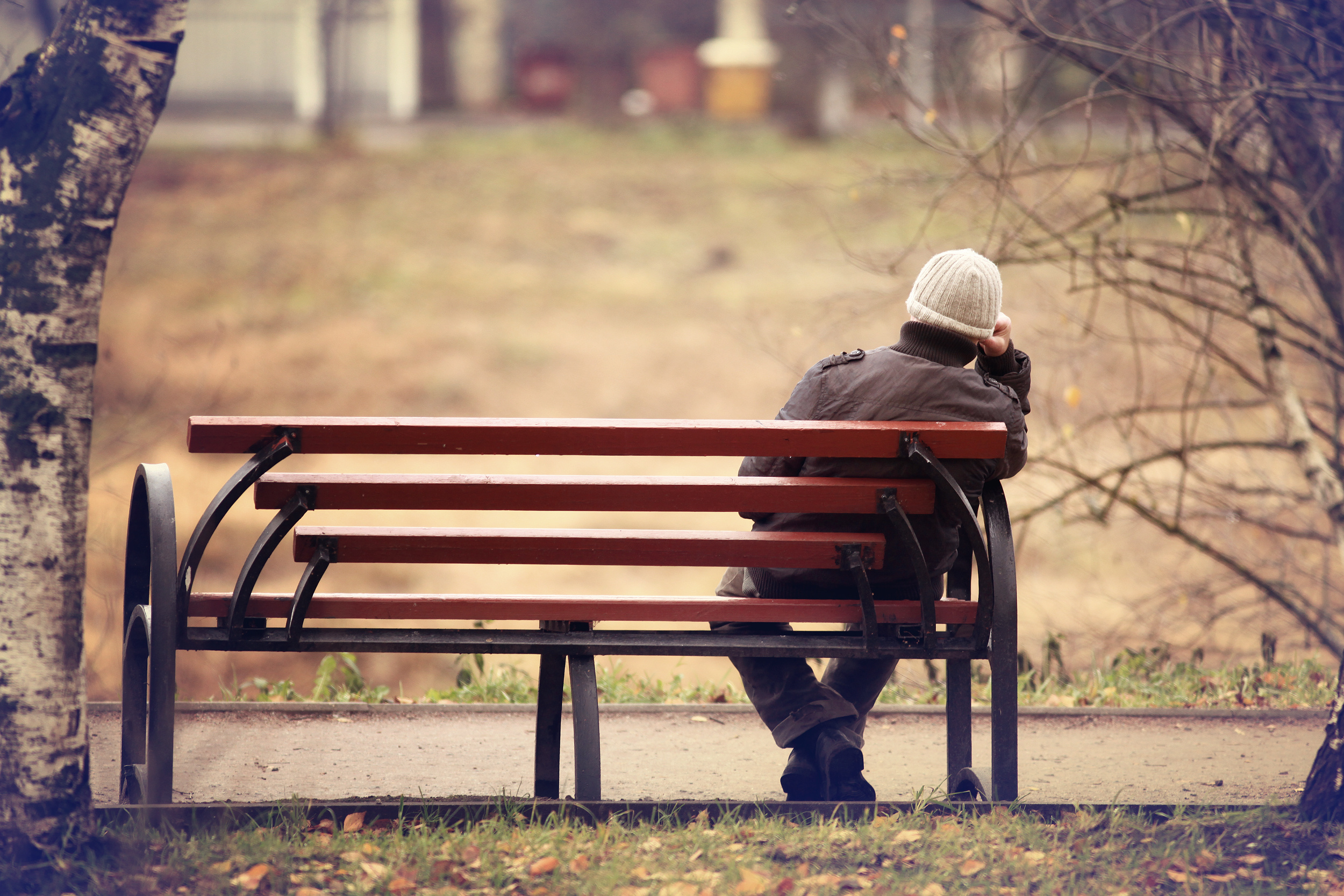 Une personne portant un bonnet est assise seule sur un banc de parc, le regard perdu dans le vide. La scène est encadrée par des troncs d'arbres avec des feuilles d'automne éparpillées sur le sol, créant une atmosphère paisible et introspective.