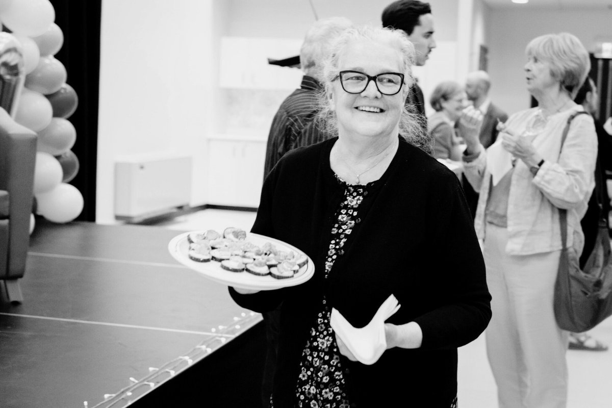 Une femme âgée avec des lunettes sourit en tenant une assiette de petites friandises lors d'une réunion sociale. Plusieurs personnes parlent à l'arrière-plan. L'image est en noir et blanc.