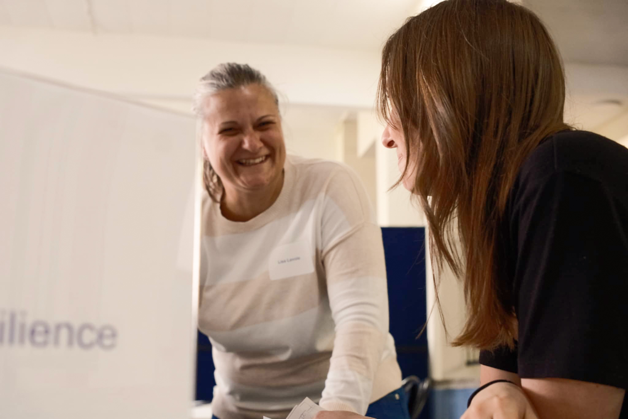 Deux femmes sourient et se parlent à l'intérieur. L'une d'entre elles fait face à l'appareil photo et rit, tandis que l'autre est tournée vers l'extérieur et engage la conversation. L'atmosphère semble amicale et détendue.