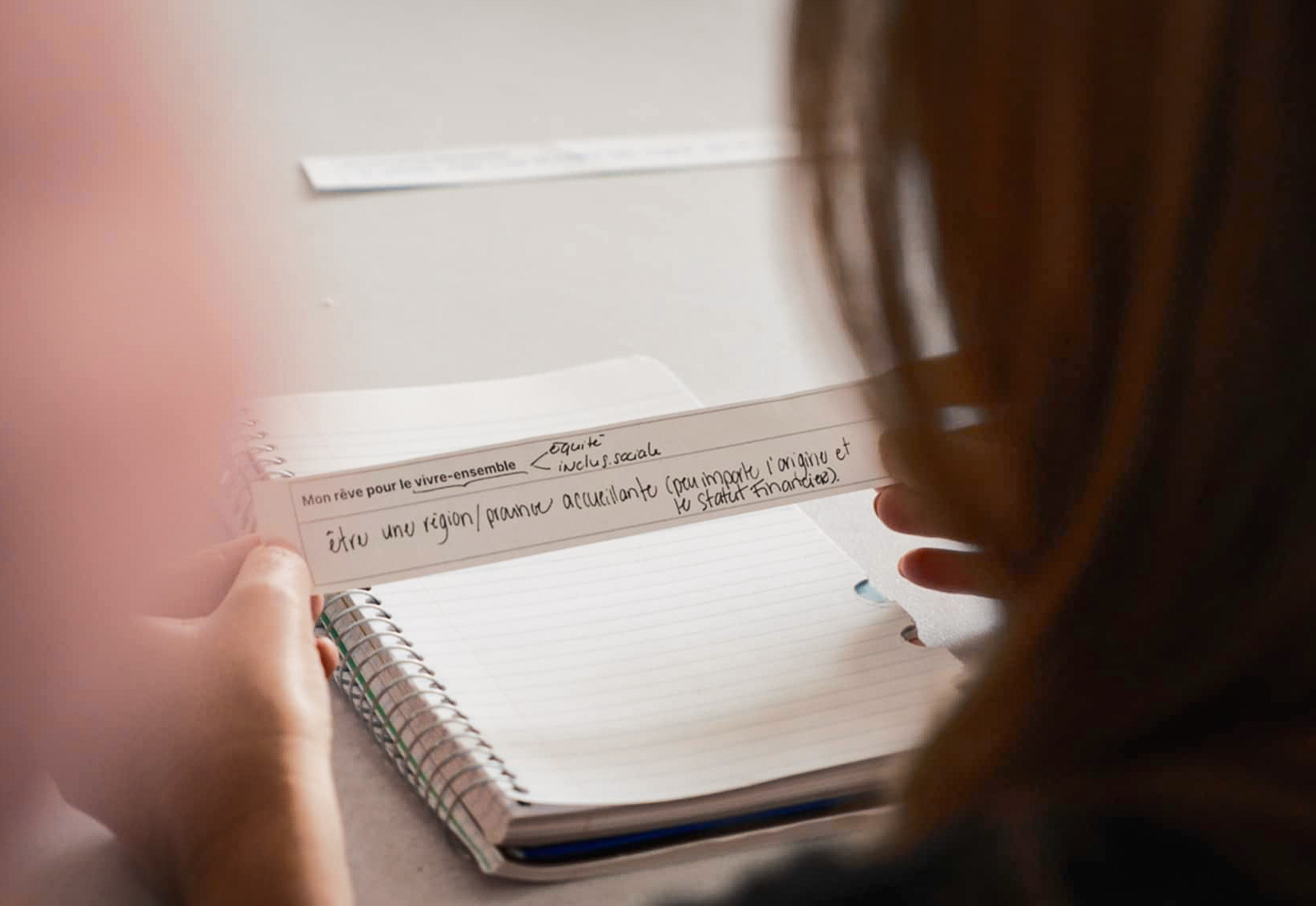 Une personne tient une note manuscrite en français au-dessus d'un carnet à spirales ouvert, une autre personne étant assise à proximité. L'accent est mis sur la note et l'arrière-plan est légèrement flou.
