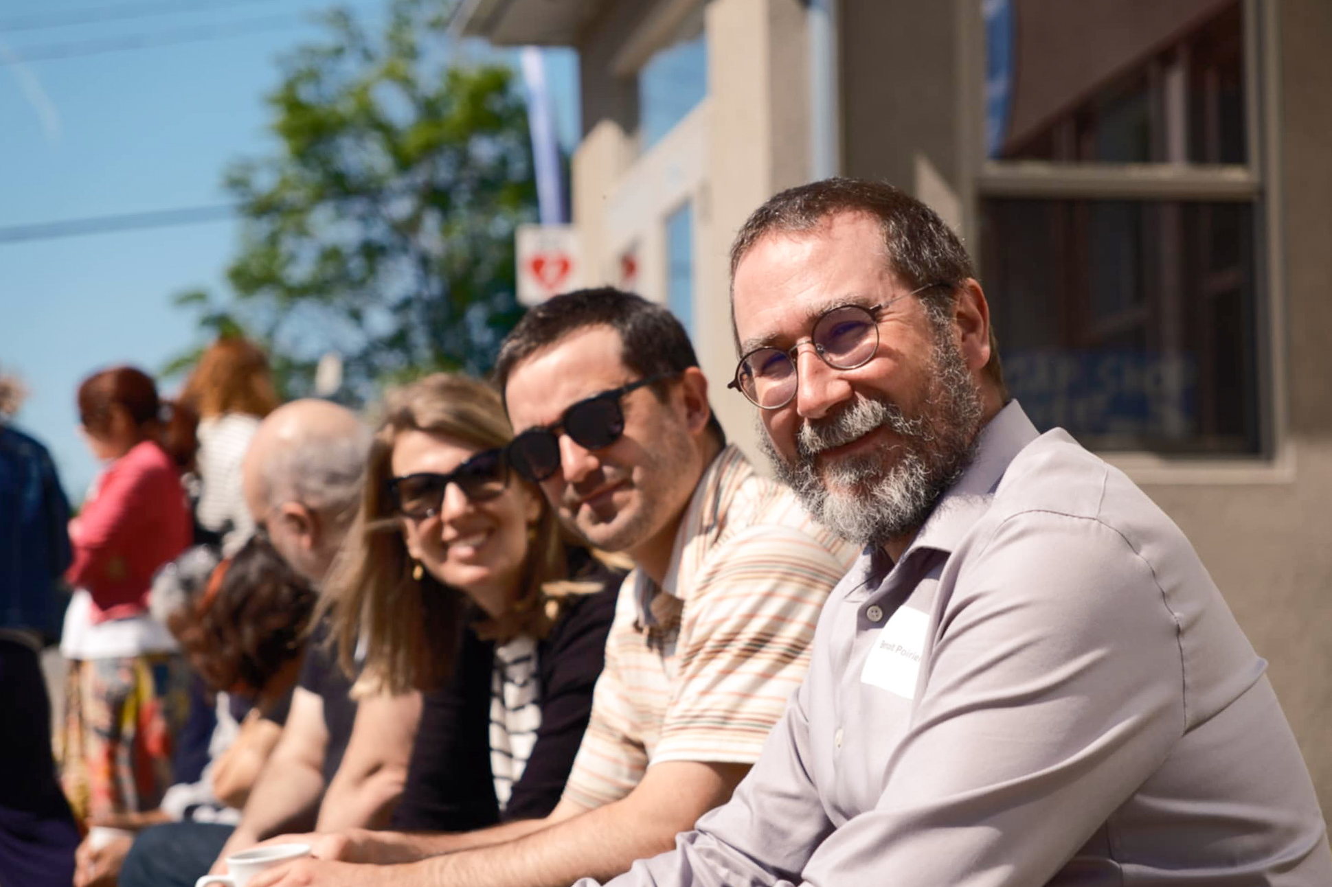 Un groupe de personnes est assis à l'extérieur, au soleil, et sourit à l'appareil photo. L'accent est mis sur un homme barbu portant des lunettes et une chemise grise, avec deux autres personnes à ses côtés portant des lunettes de soleil. Un bâtiment et des arbres se trouvent à l'arrière-plan.