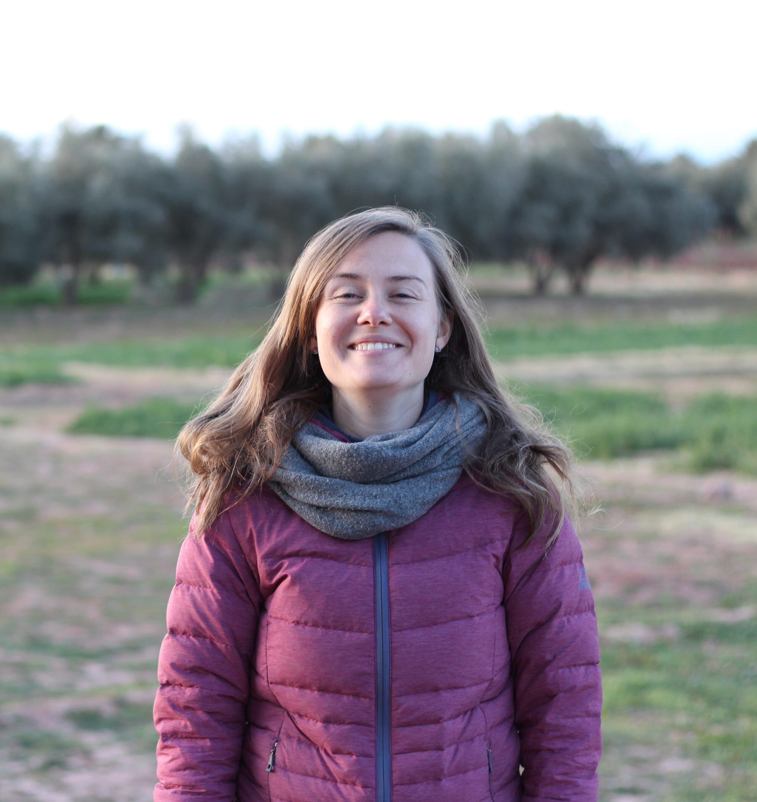Une femme aux longs cheveux bruns, portant une veste bordeaux et une écharpe grise, se tient en plein air sur un terrain herbeux avec des arbres en arrière-plan, souriant à l'appareil photo.