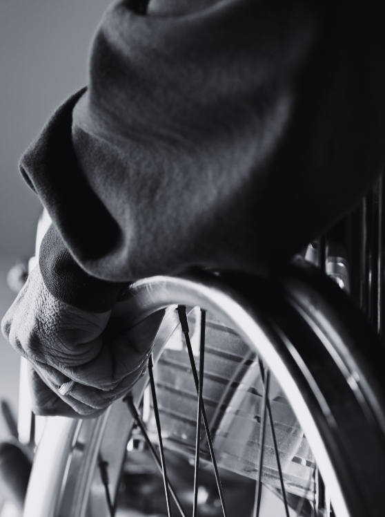 Image en gros plan, en noir et blanc, de la main d'une personne saisissant la roue d'un fauteuil roulant, mettant en évidence les rayons et une partie de l'accoudoir.