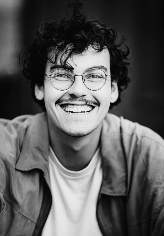 Un jeune homme aux cheveux frisés, aux lunettes et à la moustache sourit largement, vêtu d'un tee-shirt et d'une veste de couleur claire. Le portrait est en noir et blanc avec un arrière-plan flou.